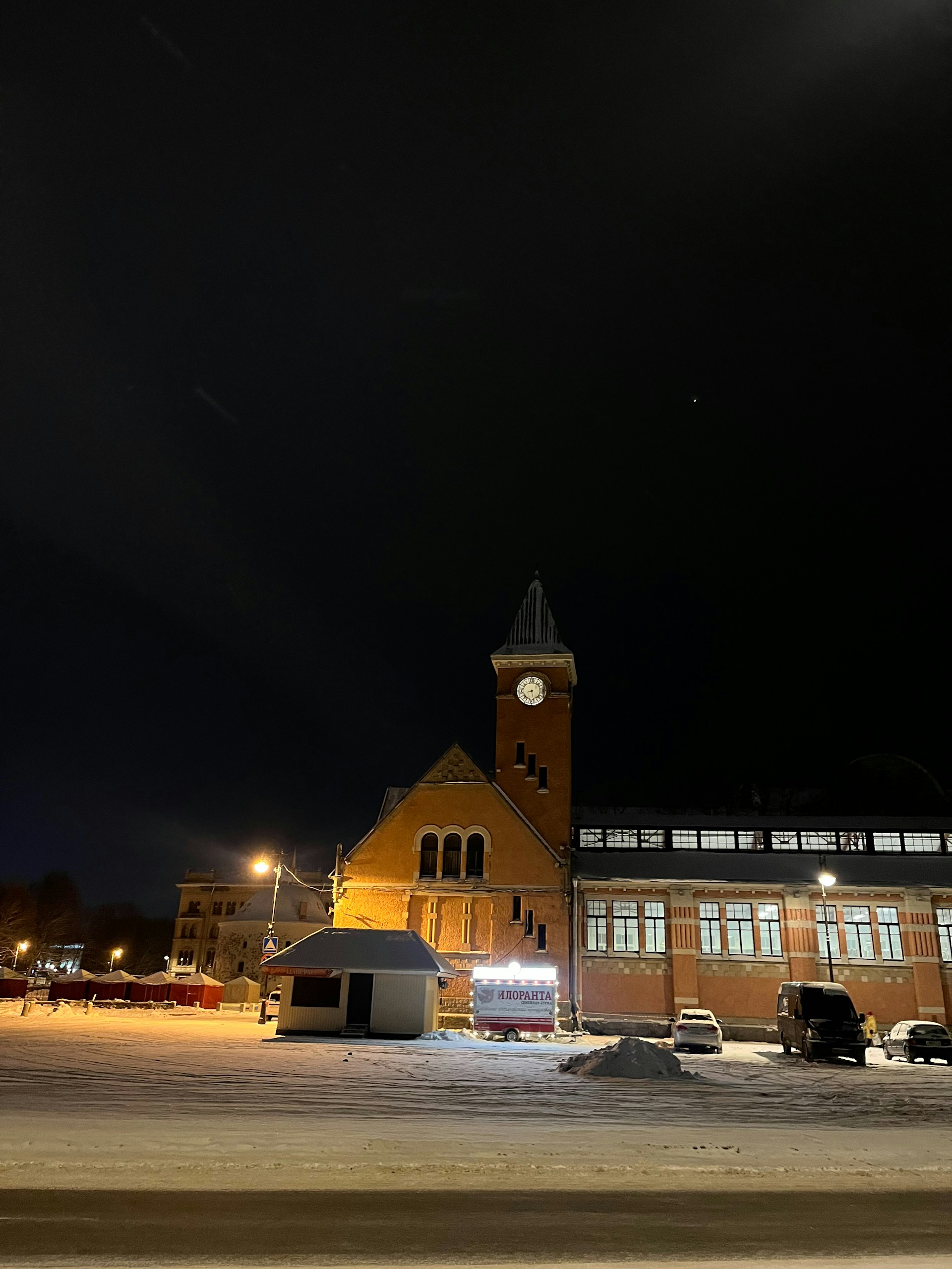 A nighttime scene features a building with a prominent clock tower and arched windows. The building has a brick facade and is illuminated by street lights. There are snow-covered grounds in the foreground, several parked vehicles, and a small structure with Russian text on it. Other buildings are visible in the background.