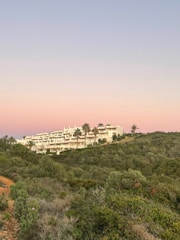 A scenic view of a modern, sprawling white building complex nestled on a hillside. It is surrounded by lush greenery and various trees, including palm trees, under a serene sky with shades of pink and blue. The natural landscape is prominent with a foreground of dense shrubbery and a dirt path.