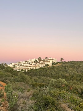 A scenic view of a modern, sprawling white building complex nestled on a hillside. It is surrounded by lush greenery and various trees, including palm trees, under a serene sky with shades of pink and blue. The natural landscape is prominent with a foreground of dense shrubbery and a dirt path.
