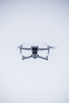 A quadcopter drone hovers in the air against a minimalistic white background. The drone is facing the camera, with its rotors visible in motion. It has a sleek, modern design and subtle LED lights near the propellers.