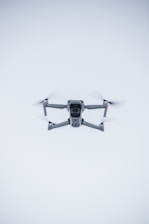 A quadcopter drone hovers in the air against a minimalistic white background. The drone is facing the camera, with its rotors visible in motion. It has a sleek, modern design and subtle LED lights near the propellers.