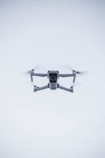 A quadcopter drone hovers in the air against a minimalistic white background. The drone is facing the camera, with its rotors visible in motion. It has a sleek, modern design and subtle LED lights near the propellers.