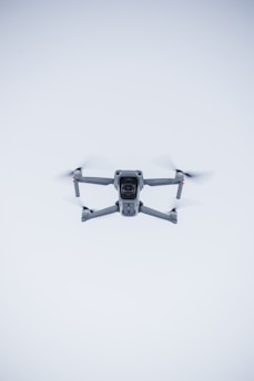 A quadcopter drone hovers in the air against a minimalistic white background. The drone is facing the camera, with its rotors visible in motion. It has a sleek, modern design and subtle LED lights near the propellers.