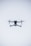 A quadcopter drone hovers in the air against a minimalistic white background. The drone is facing the camera, with its rotors visible in motion. It has a sleek, modern design and subtle LED lights near the propellers.