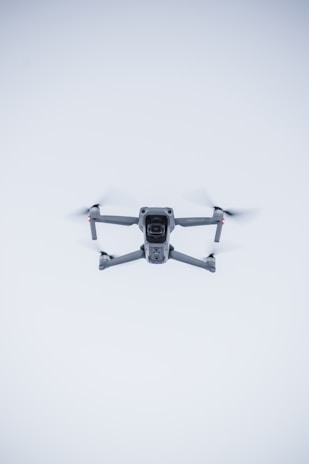 A quadcopter drone hovers in the air against a minimalistic white background. The drone is facing the camera, with its rotors visible in motion. It has a sleek, modern design and subtle LED lights near the propellers.