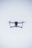 A quadcopter drone hovers in the air against a minimalistic white background. The drone is facing the camera, with its rotors visible in motion. It has a sleek, modern design and subtle LED lights near the propellers.
