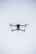 A quadcopter drone hovers in the air against a minimalistic white background. The drone is facing the camera, with its rotors visible in motion. It has a sleek, modern design and subtle LED lights near the propellers.