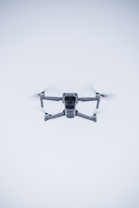 A quadcopter drone hovers in the air against a minimalistic white background. The drone is facing the camera, with its rotors visible in motion. It has a sleek, modern design and subtle LED lights near the propellers.