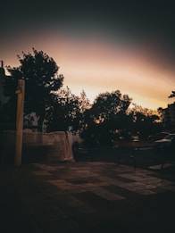 A serene outdoor scene during twilight. The silhouettes of trees are dark against a gradient sky transitioning from orange to deep blue. The foreground features a tiled pavement with subdued lighting, and some covered objects are visible, suggesting urban elements.
