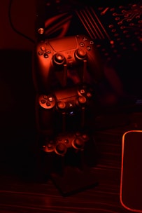 A stack of gaming controllers is illuminated by red lighting, creating a moody atmosphere. The controllers are placed on a stand, and the surrounding environment hints at a tech setup with circuit-like designs in the background.