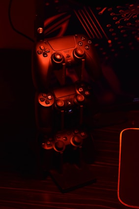 A stack of gaming controllers is illuminated by red lighting, creating a moody atmosphere. The controllers are placed on a stand, and the surrounding environment hints at a tech setup with circuit-like designs in the background.