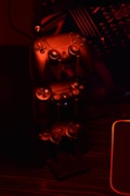 A stack of gaming controllers is illuminated by red lighting, creating a moody atmosphere. The controllers are placed on a stand, and the surrounding environment hints at a tech setup with circuit-like designs in the background.