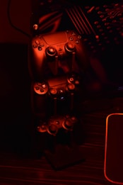 A stack of gaming controllers is illuminated by red lighting, creating a moody atmosphere. The controllers are placed on a stand, and the surrounding environment hints at a tech setup with circuit-like designs in the background.