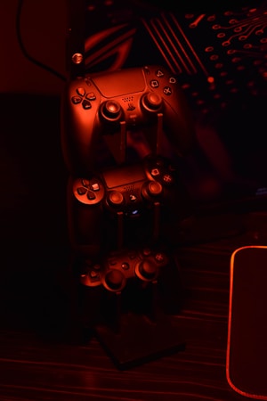 A stack of gaming controllers is illuminated by red lighting, creating a moody atmosphere. The controllers are placed on a stand, and the surrounding environment hints at a tech setup with circuit-like designs in the background.
