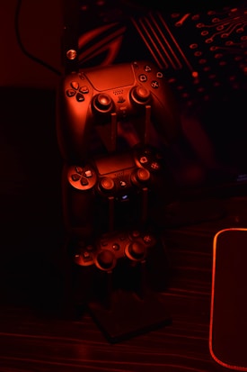 A stack of gaming controllers is illuminated by red lighting, creating a moody atmosphere. The controllers are placed on a stand, and the surrounding environment hints at a tech setup with circuit-like designs in the background.
