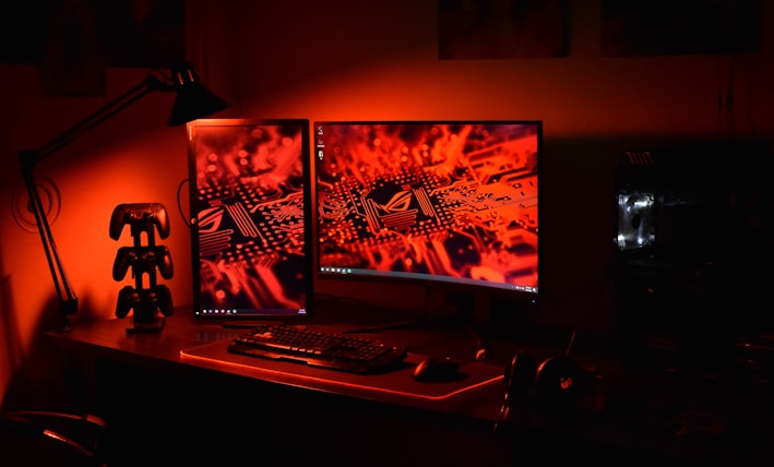 A computer desk setup is illuminated in red light. Two monitors display an electronic circuitry pattern. The desk holds a keyboard, mouse, and a game controller stand. An adjustable desk lamp is positioned to the side, and a gaming PC with visible internal lighting sits to the right.