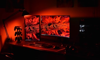 A computer desk setup is illuminated in red light. Two monitors display an electronic circuitry pattern. The desk holds a keyboard, mouse, and a game controller stand. An adjustable desk lamp is positioned to the side, and a gaming PC with visible internal lighting sits to the right.