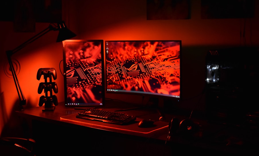 A computer desk setup is illuminated in red light. Two monitors display an electronic circuitry pattern. The desk holds a keyboard, mouse, and a game controller stand. An adjustable desk lamp is positioned to the side, and a gaming PC with visible internal lighting sits to the right.