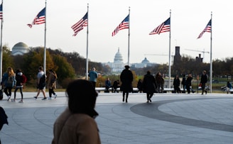 A group of people walking and standing near a paved area adorned with American flags on tall flagpoles. The background features an iconic white dome building, presumably a government structure, with a park-like setting and trees showcasing autumn colors. There are also several other buildings and construction cranes visible in the distance.