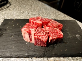 Several pieces of raw, marbled beef are neatly arranged on a flat, black slate serving tray. The beef is seasoned with ground black pepper and the background consists of a speckled gray surface, possibly a countertop.