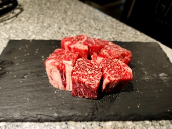 Several pieces of raw, marbled beef are neatly arranged on a flat, black slate serving tray. The beef is seasoned with ground black pepper and the background consists of a speckled gray surface, possibly a countertop.