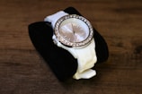 A white analog wristwatch with a band placed on a black cushion. The watch features a round face adorned with small rhinestones around the bezel, giving it an elegant and luxurious appearance. The background is a wooden surface, enhancing the watch's sophisticated style.