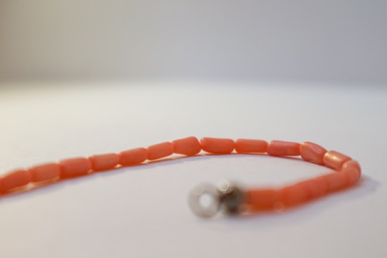 A beaded bracelet or necklace with small, coral-colored beads arranged in a single string pattern. The beads have an elongated, tubular shape with a circular clasp attached at one end. The background is plain and softly lit, creating a minimalistic and focused presentation of the jewelry piece.