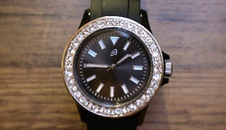 A stylish wristwatch with a black face and a silver frame adorned with sparkling stones around the perimeter. The watch has a black band and a silver crown on the right side. The face features simple, rectangular hour markers and two hands indicating the time.