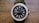 A stylish wristwatch with a black face and a silver frame adorned with sparkling stones around the perimeter. The watch has a black band and a silver crown on the right side. The face features simple, rectangular hour markers and two hands indicating the time.