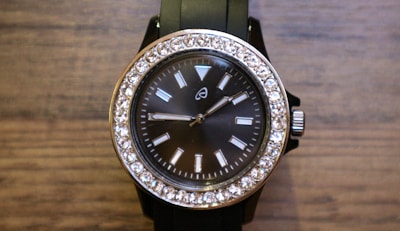 A stylish wristwatch with a black face and a silver frame adorned with sparkling stones around the perimeter. The watch has a black band and a silver crown on the right side. The face features simple, rectangular hour markers and two hands indicating the time.