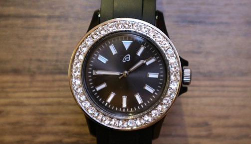A stylish wristwatch with a black face and a silver frame adorned with sparkling stones around the perimeter. The watch has a black band and a silver crown on the right side. The face features simple, rectangular hour markers and two hands indicating the time.