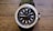 A stylish wristwatch with a black face and a silver frame adorned with sparkling stones around the perimeter. The watch has a black band and a silver crown on the right side. The face features simple, rectangular hour markers and two hands indicating the time.