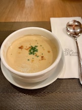 A creamy soup served in a white bowl on a matching white saucer, garnished with finely chopped parsley and croutons. It is placed on a brown woven placemat, next to a folded napkin with a spoon on top of it.