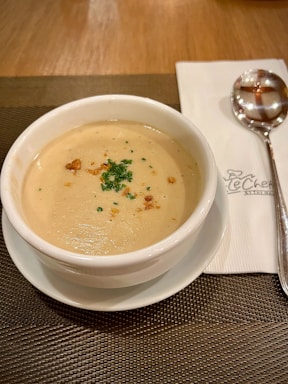 A creamy soup served in a white bowl on a matching white saucer, garnished with finely chopped parsley and croutons. It is placed on a brown woven placemat, next to a folded napkin with a spoon on top of it.