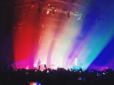 A colorful concert scene filled with vibrant lighting effects. Silhouetted people in the audience hold up phones, capturing the event. The stage is highlighted by diagonal beams of red, orange, yellow, green, blue, and purple light, which cascade over the crowd, creating a dynamic and energetic atmosphere.