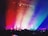 A colorful concert scene filled with vibrant lighting effects. Silhouetted people in the audience hold up phones, capturing the event. The stage is highlighted by diagonal beams of red, orange, yellow, green, blue, and purple light, which cascade over the crowd, creating a dynamic and energetic atmosphere.
