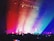 A colorful concert scene filled with vibrant lighting effects. Silhouetted people in the audience hold up phones, capturing the event. The stage is highlighted by diagonal beams of red, orange, yellow, green, blue, and purple light, which cascade over the crowd, creating a dynamic and energetic atmosphere.