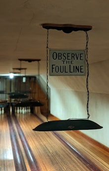 A vintage bowling alley with wooden lanes and a sign hanging that reads 'Observe the Foul Line'. The lanes are polished and lead towards a set of bowling pins in the distance. The lighting is dim, giving the scene a nostalgic feel.
