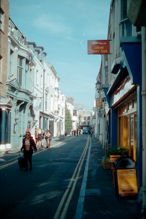 A narrow street flanked by buildings with various architectural styles, bustling with pedestrians. A sign for Indian street food is visible, and a person is pulling a suitcase along the street. The sky is clear and blue, contributing to a vibrant, urban scene.