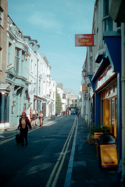 A narrow street flanked by buildings with various architectural styles, bustling with pedestrians. A sign for Indian street food is visible, and a person is pulling a suitcase along the street. The sky is clear and blue, contributing to a vibrant, urban scene.