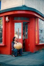 A person is sitting outside a red-painted storefront, holding and seemingly playing a large stringed musical instrument. The store appears to be related to percussion, as indicated by signage. The scene looks urban with a paved sidewalk, and the windows have a grid of small square panes.