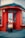 A person is sitting outside a red-painted storefront, holding and seemingly playing a large stringed musical instrument. The store appears to be related to percussion, as indicated by signage. The scene looks urban with a paved sidewalk, and the windows have a grid of small square panes.