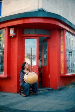 A person is sitting outside a red-painted storefront, holding and seemingly playing a large stringed musical instrument. The store appears to be related to percussion, as indicated by signage. The scene looks urban with a paved sidewalk, and the windows have a grid of small square panes.