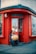 A person is sitting outside a red-painted storefront, holding and seemingly playing a large stringed musical instrument. The store appears to be related to percussion, as indicated by signage. The scene looks urban with a paved sidewalk, and the windows have a grid of small square panes.