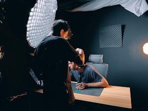 Two people are in a dark room with professional lighting equipment. One person appears to be adjusting a camera or light, while the other sits at a wooden desk with a microphone, facing the setup. Acoustic foam panels are mounted on the wall, and a bright light source is visible to the side.