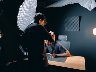 Two people are in a dark room with professional lighting equipment. One person appears to be adjusting a camera or light, while the other sits at a wooden desk with a microphone, facing the setup. Acoustic foam panels are mounted on the wall, and a bright light source is visible to the side.