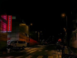A quiet, dimly-lit street at night with an illuminated hospital emergency sign on the left side of the image. The street is mostly empty, featuring a white van parked near the side, and streetlights casting a soft glow on the wet pavement. The surrounding buildings appear mostly dark, adding to the nighttime ambiance.