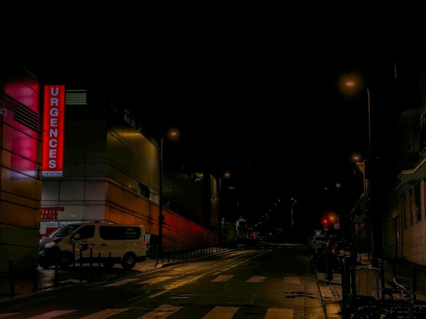 A quiet, dimly-lit street at night with an illuminated hospital emergency sign on the left side of the image. The street is mostly empty, featuring a white van parked near the side, and streetlights casting a soft glow on the wet pavement. The surrounding buildings appear mostly dark, adding to the nighttime ambiance.