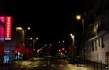 A nighttime urban street scene featuring an illuminated emergency sign with the word 'Urgences' in bright red letters on a building. Streetlights cast a warm glow over the wet pavement and the surrounding buildings are mostly in shadow. The street is empty, adding a sense of quiet and stillness.