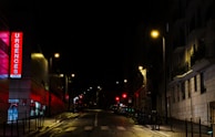 A nighttime urban street scene featuring an illuminated emergency sign with the word 'Urgences' in bright red letters on a building. Streetlights cast a warm glow over the wet pavement and the surrounding buildings are mostly in shadow. The street is empty, adding a sense of quiet and stillness.